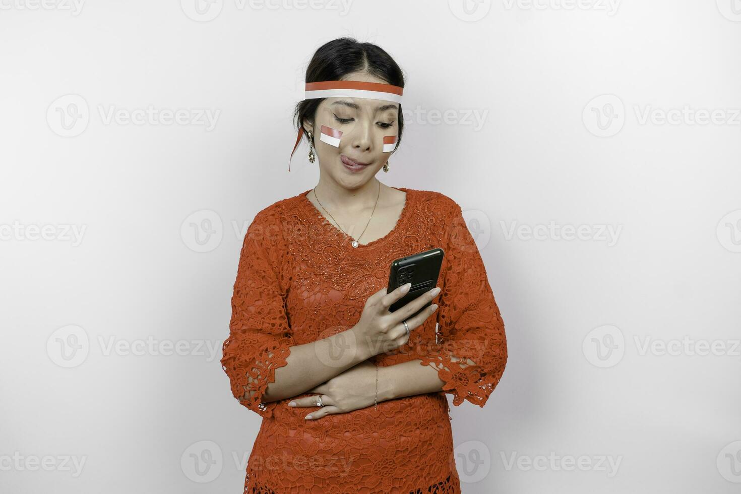un pensativo asiático mujer vistiendo rojo kebaya y venda, participación su teléfono y estómago, pensando acerca de qué a comer o orden, aislado por blanco antecedentes. de indonesia independencia día foto