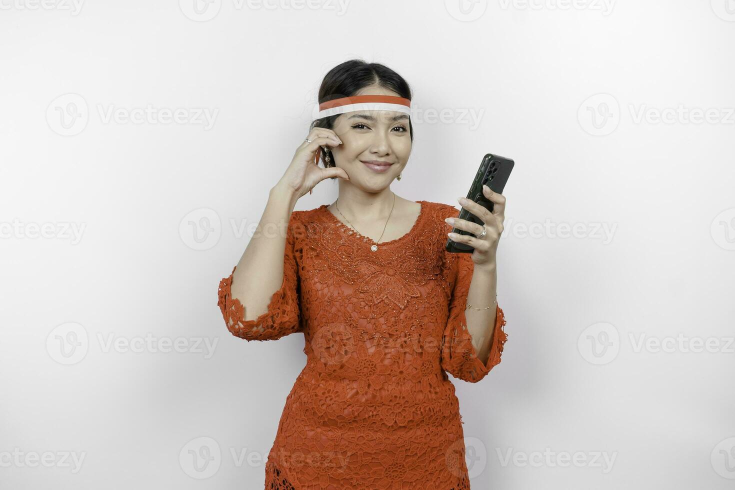 A happy Asian woman wearing red kebaya and headband, holding her phone, and gesturing love sign by her fingers showing tender feeling, isolated by white background. Indonesia's independence day photo