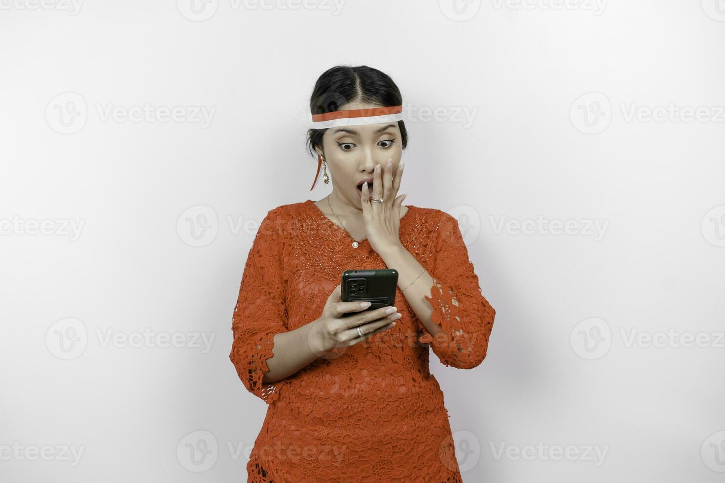 A portrait of a shocked Asian woman wearing red kebaya and headband, holding her phone while her mouth wide open, isolated by white background. Indonesia's independence day concept photo