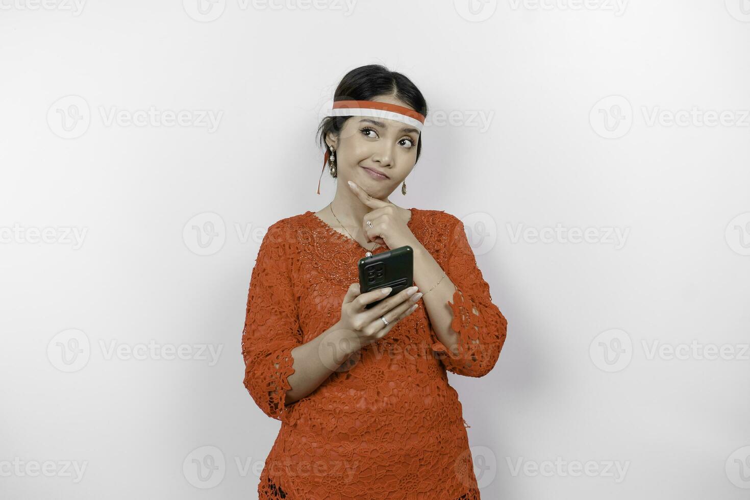 A smart young woman wearing headband and red kebaya is looking aside to an idea on copy space while holding her phone, isolated by white background. Indonesia's independence day. photo