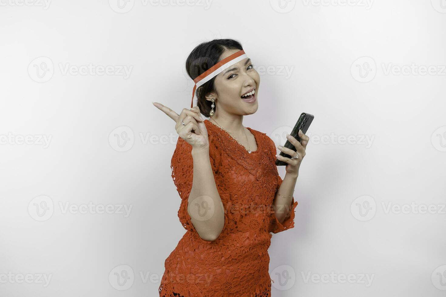 A happy Asian woman wearing red kebaya and headband, holding her phone, and pointing copy space on top of her, isolated by white background. Indonesia's independence day photo