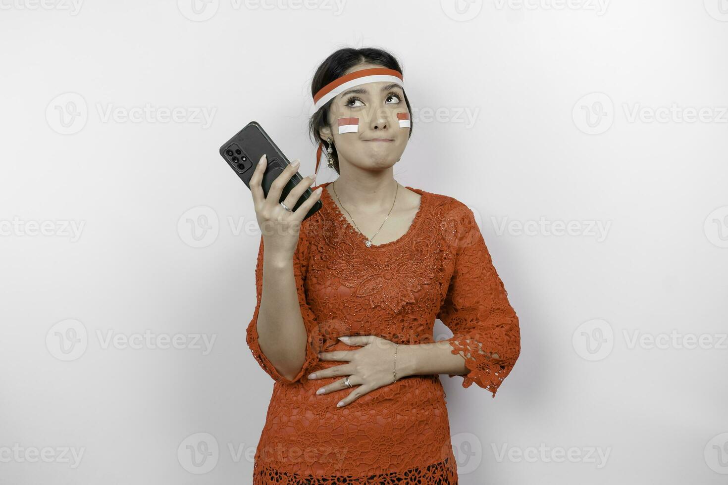 A thoughtful Asian woman wearing red kebaya and headband, holding her phone and stomach, thinking about what to eat or order, isolated by white background. Indonesia's independence day photo