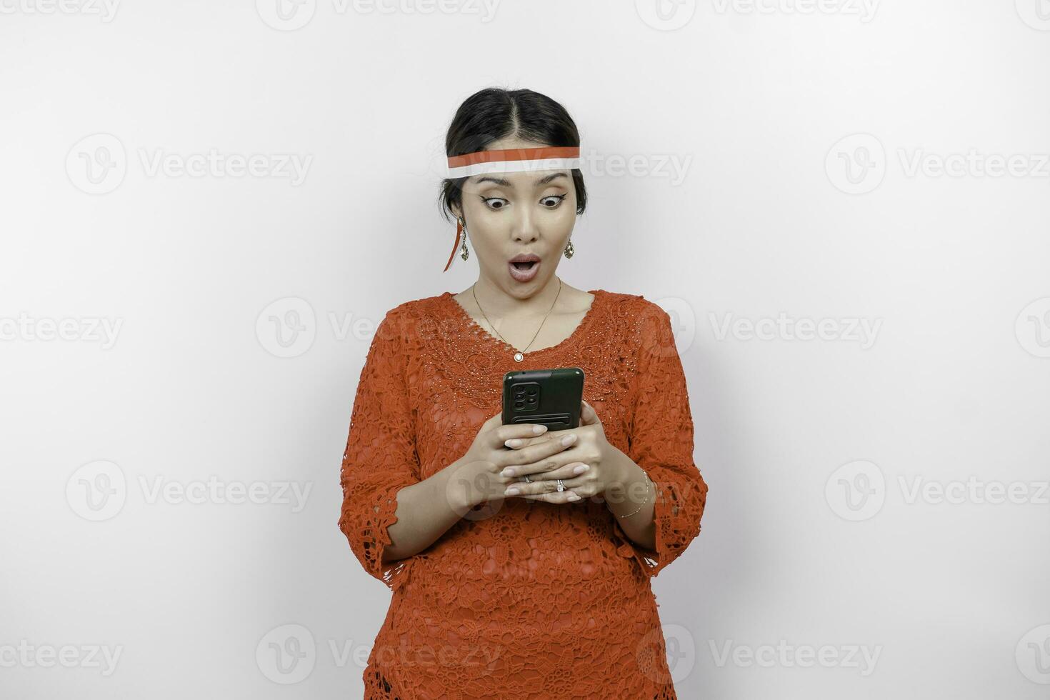 A portrait of a shocked Asian woman wearing red kebaya and headband, holding her phone while her mouth wide open, isolated by white background. Indonesia's independence day concept photo