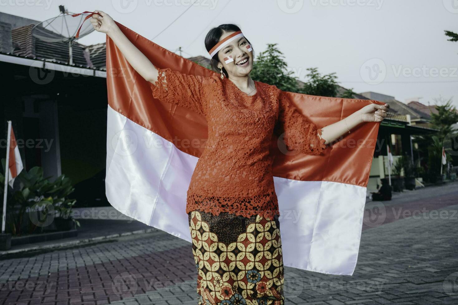 Happy smiling Indonesian woman wearing red kebaya holding Indonesia's flag to celebrate Indonesia Independence Day. Outdoor photoshoot concept photo