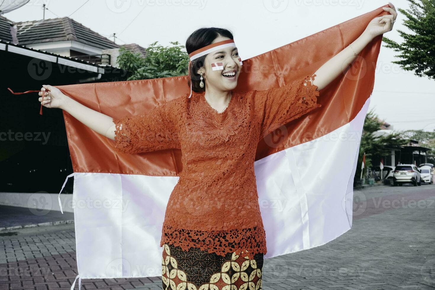 Happy smiling Indonesian woman wearing red kebaya holding Indonesia's flag to celebrate Indonesia Independence Day. Outdoor photoshoot concept photo