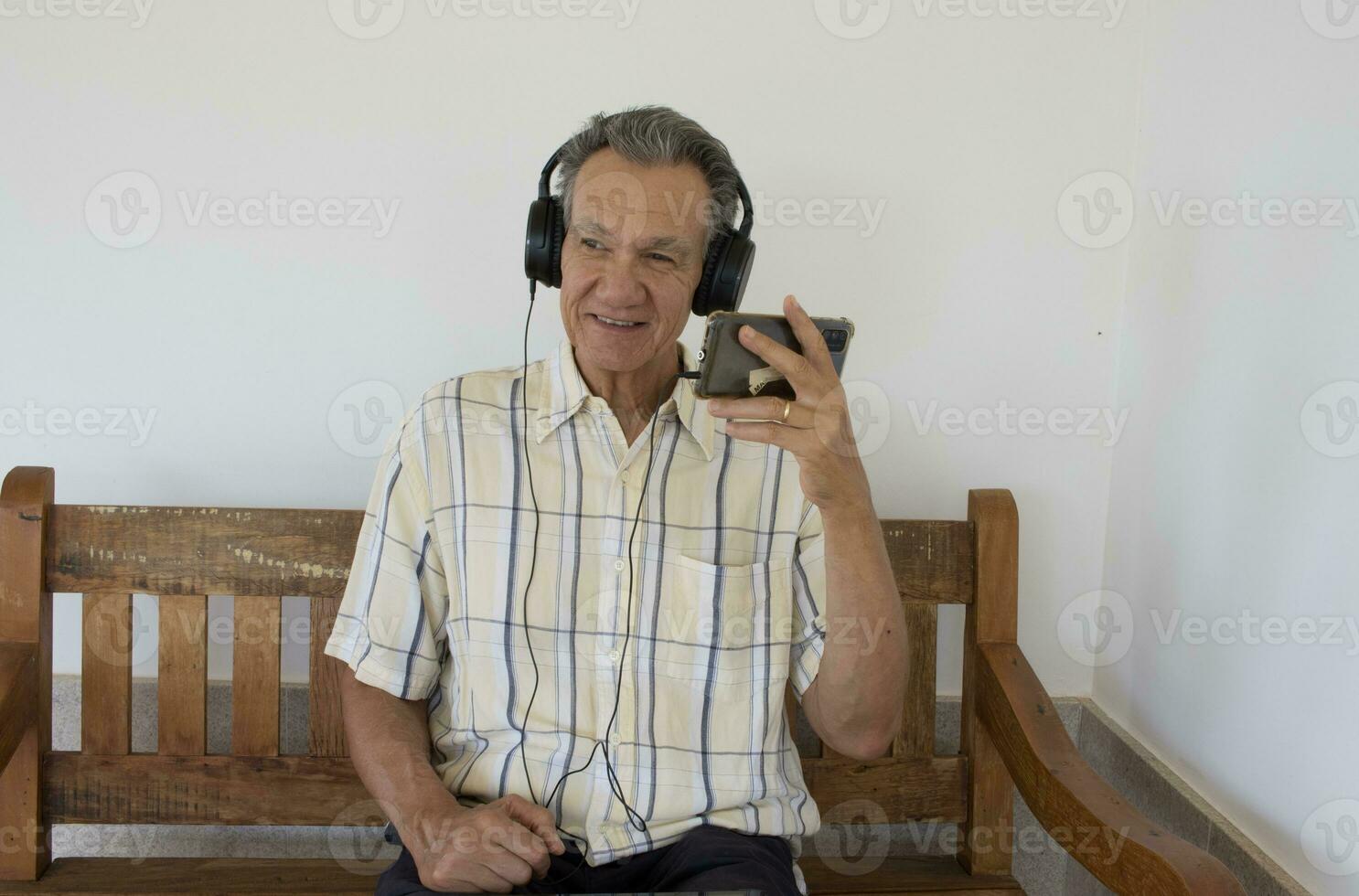 Mature Man outside sitting on a bench watching a show on his Smart Phone wearing headphones photo