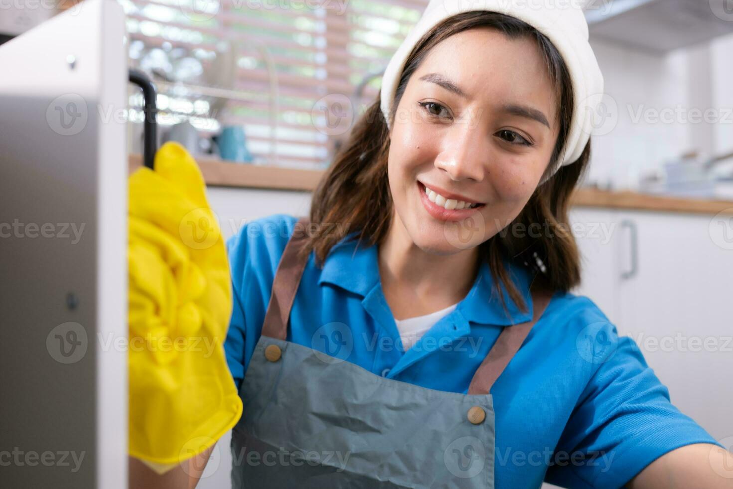 Smiling young woman in apron and cleaning gloves open kitchen cabinets photo