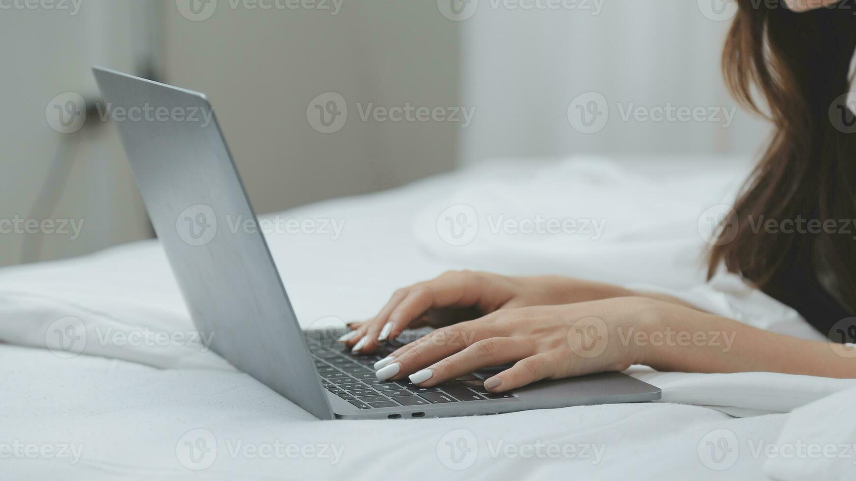 Happy young female in casual clothes smiling and looking at camera while using laptop on bed in bedroom at home photo