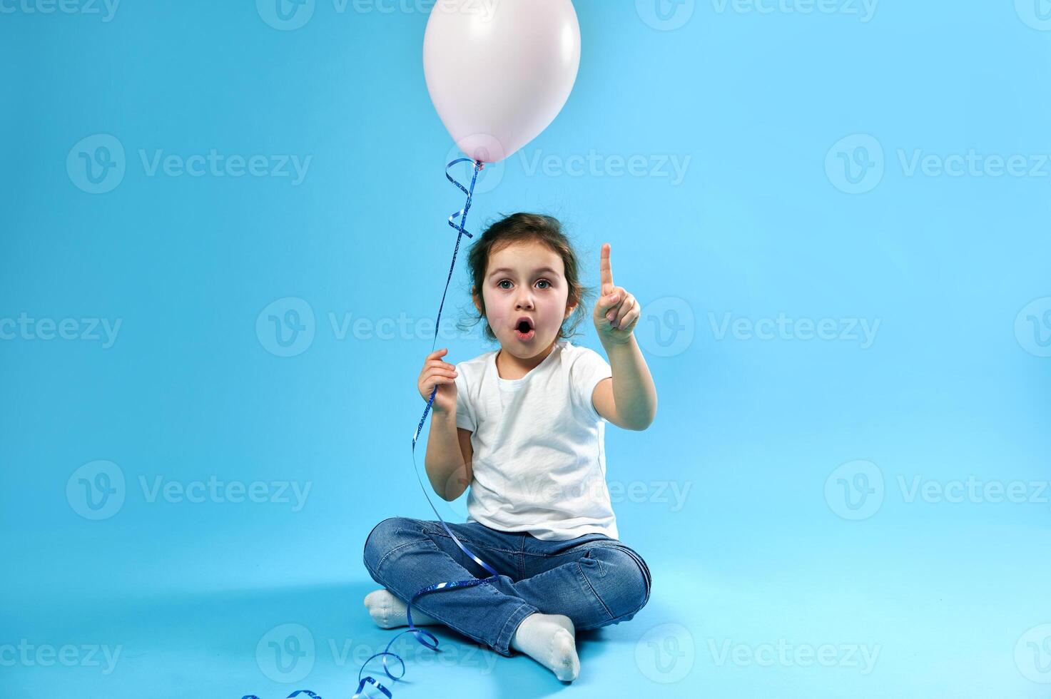 Adorable little girl with a pink balloon in her hand pointing her finger up on a blue background with copy space for promotion and advertising. photo