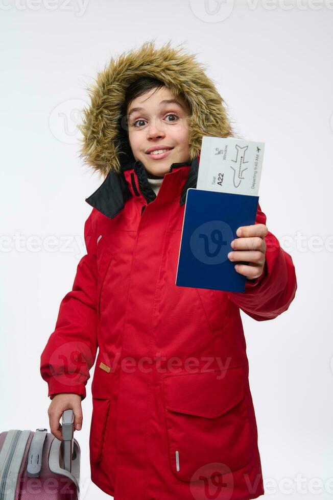 Beautiful preadolescent child, Caucasian boy in bright red parka with hood looking at camera showing a travel passport with boarding pass, isolated on white background with copy space for ads photo