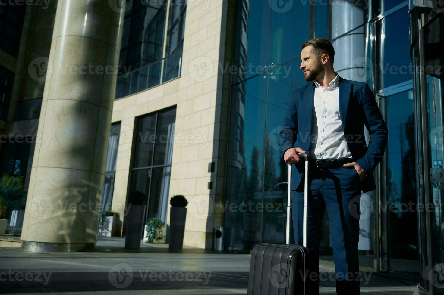 Full-length portrait of confident businessman in casual business suit standing with black suitcase at the glass entrance of the hotel, looking confidently to the side, holding his hand in his pocket photo