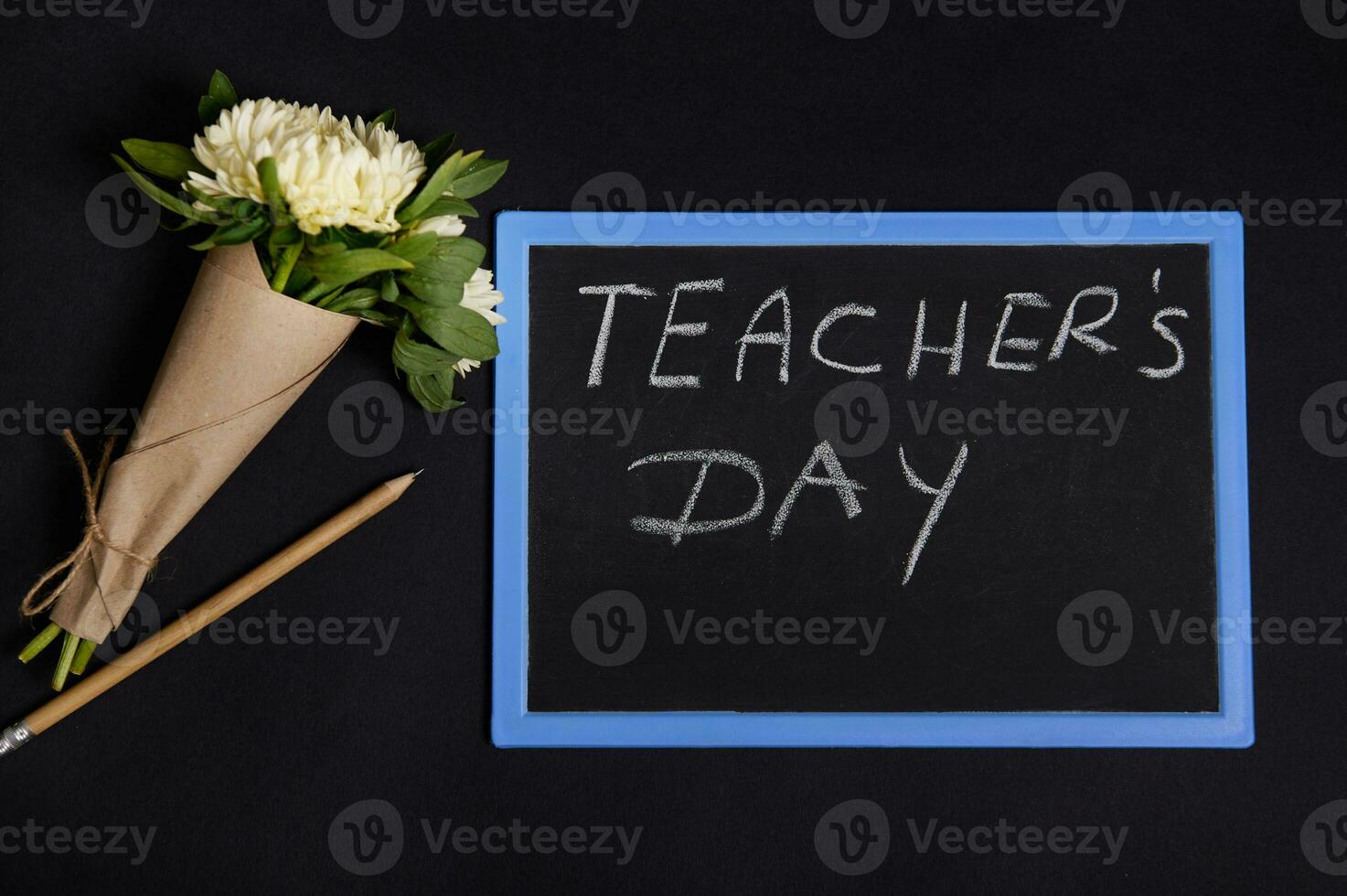 Flat lay of wooden pencil and beautiful bouquet of asters flowers in craft wrapping paper lying down next to a chalk board with lettering Teacher's Day, isolated on black background with copy space photo