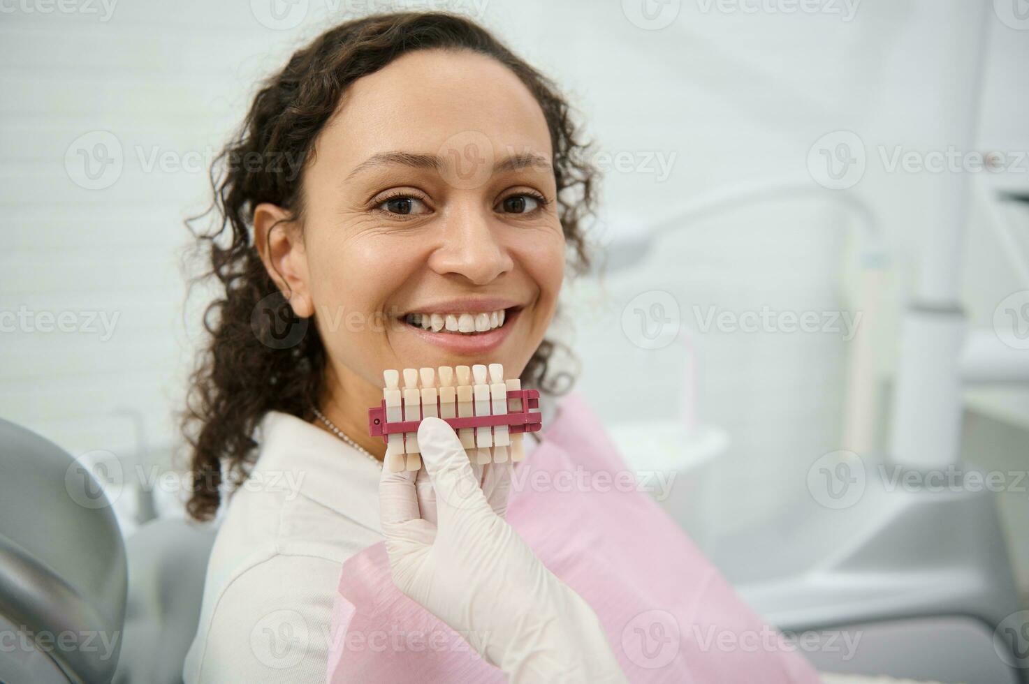 Close-up of dentist hand holding a teeth color chart near the face of a dentistry clinic woman patient smiling with beautiful toothy smile looking at camera. Teeth whitening, bleaching concept photo