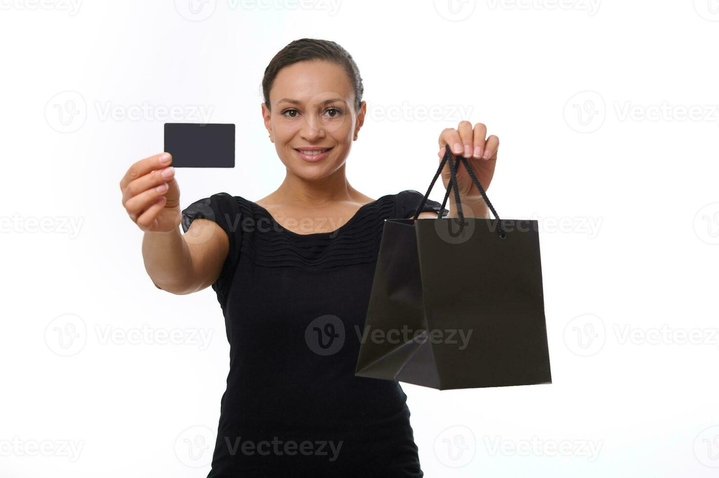 Beautiful mixed race woman looks at camera and smiles toothy smile, showing black shopping packet and plastic credit or discount card in her outstretched hands. Concept of Black Friday with copy space photo