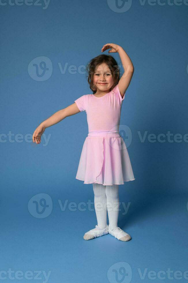 A cute smiling ballerina in pink dress performs ballet dance, standing on blue background with copy space photo