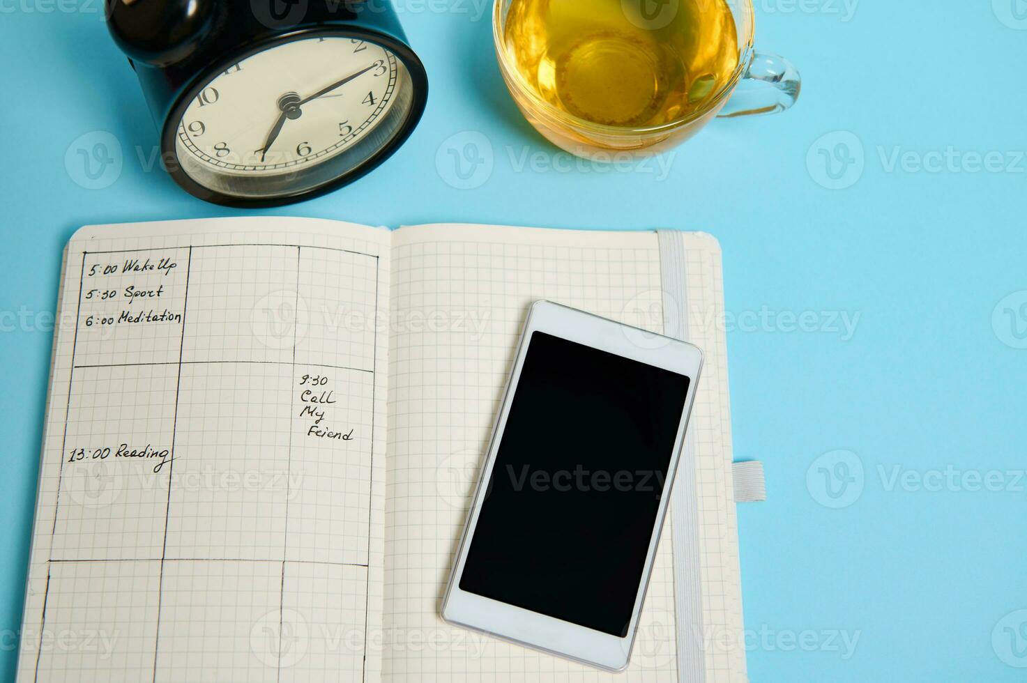 Flat lay of open notebook with schedule, smartphone, glass cup of tea and black alarm clock lie on blue surface. Color background with copy space for text. Time management, deadline, schedule concept photo