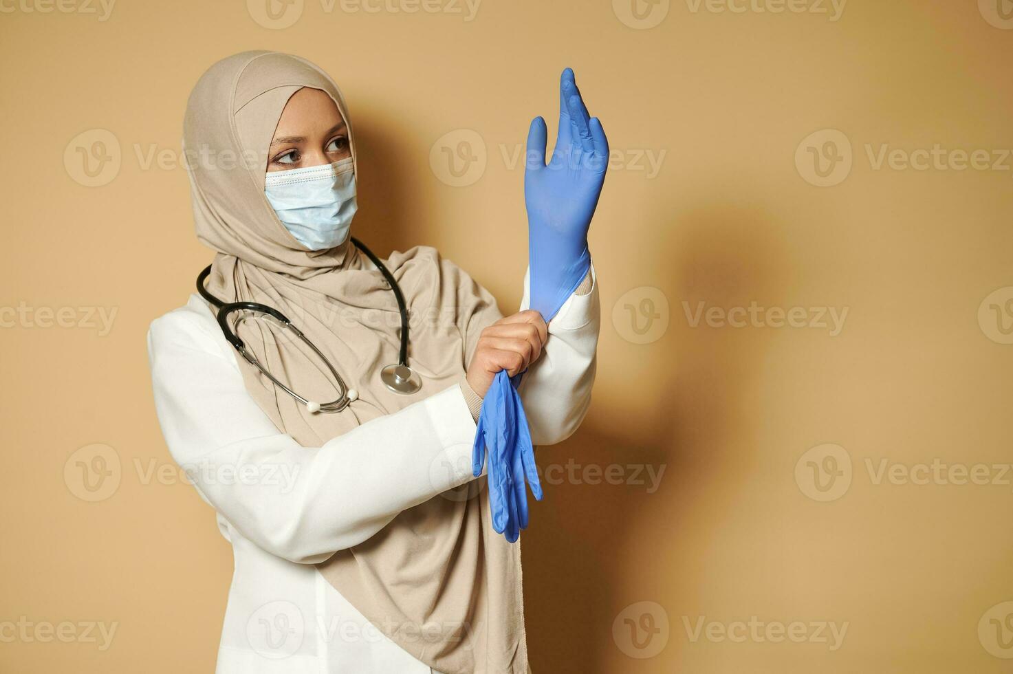 Muslim female doctor with covered head wearing medical protective gloves posing in front of camera on beige background with copy space photo