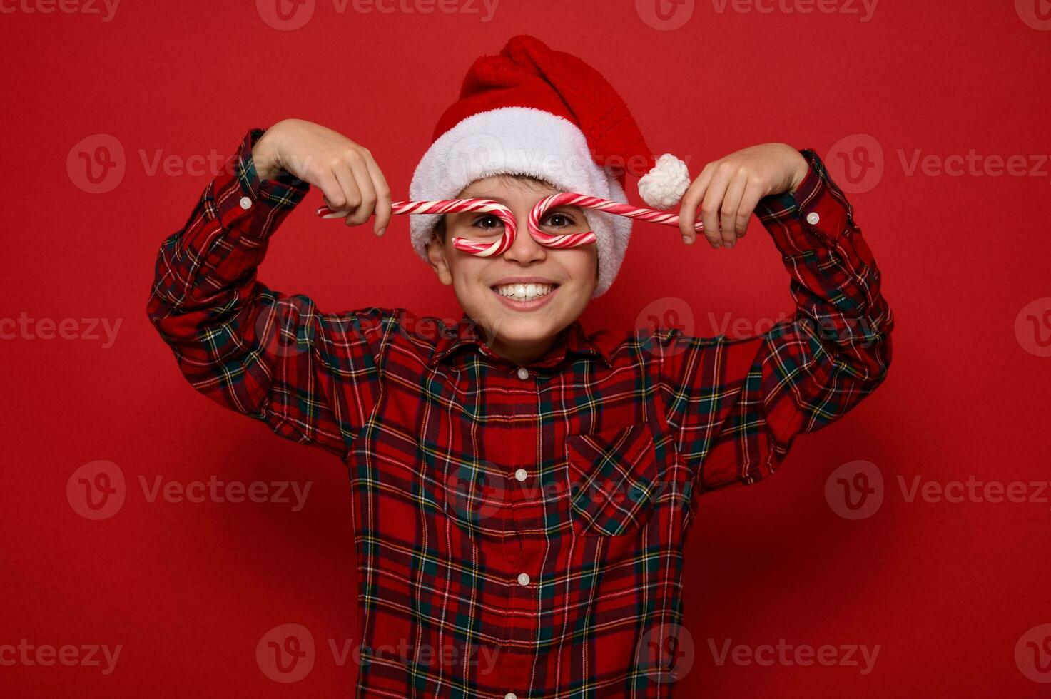 hermoso niño chico en Papa Noel claus sombrero y tartán camisa sonrisas mirando a cámara mediante Navidad dulce caramelo bastones, participación ellos imitando los anteojos, posando en rojo antecedentes con Copiar espacio para anuncio foto