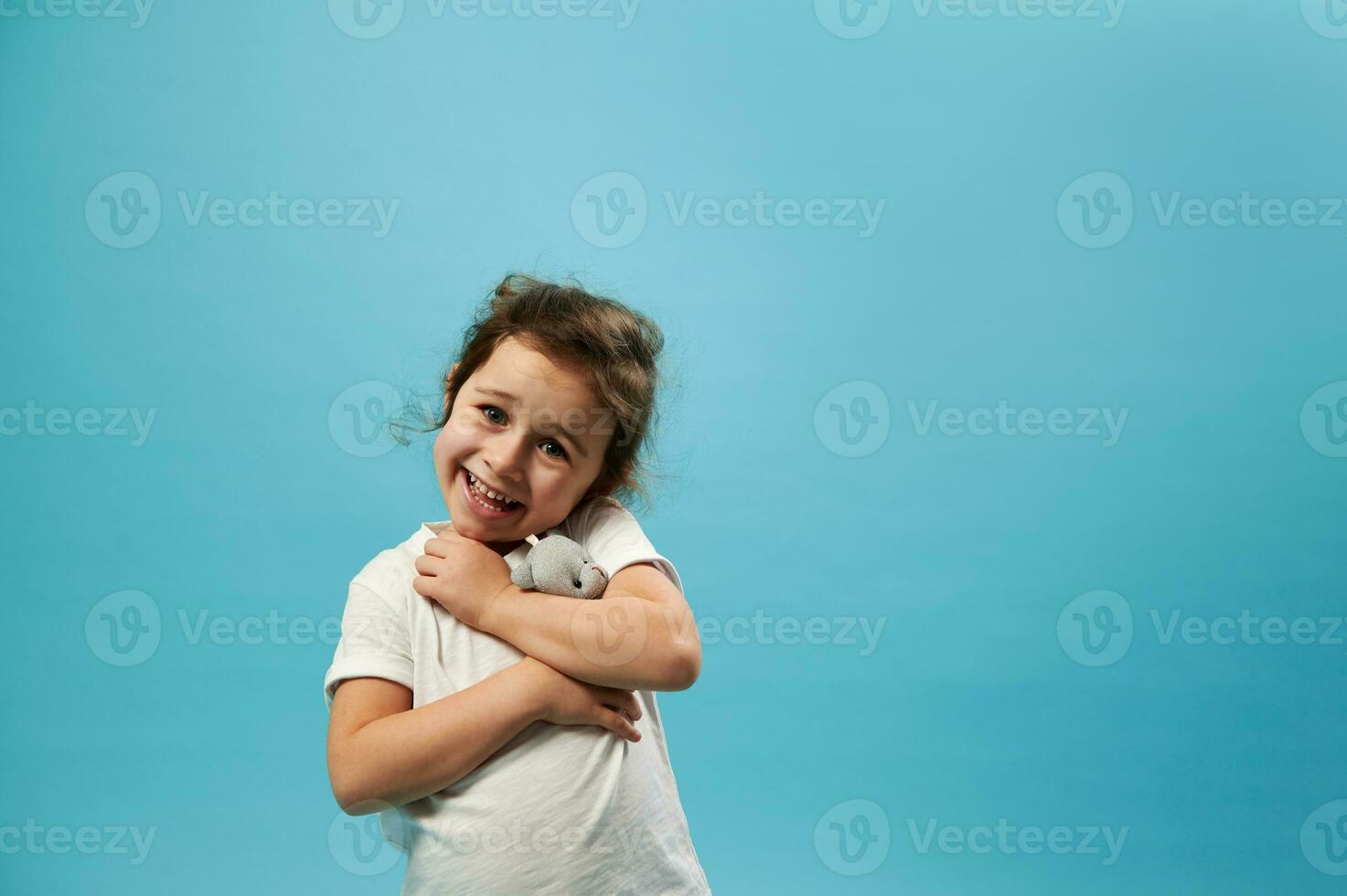 Adorable girl gently hugs her soft toy bear and looks at the camera, standing on a blue background with copy space photo
