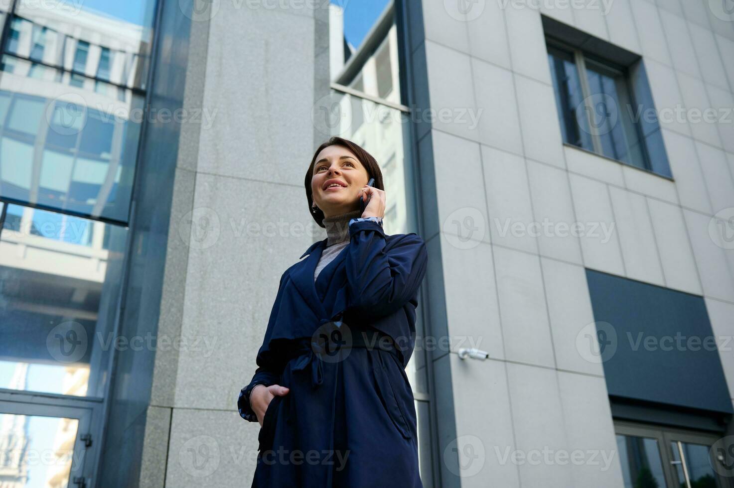 Bottom view of an attractive freelancer woman, business person, entrepreneur talking on a mobile phone on the background of urban modern corporate high-rise buildings photo