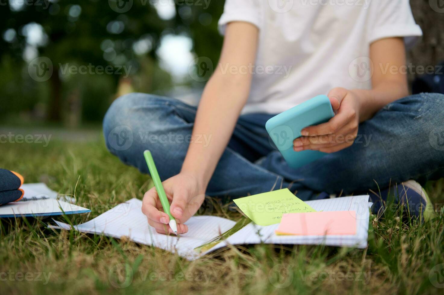 Cropped image of an unrecogniable school boy studying in the park, sitting on green grass and solving mathematics task, using smartphone and mobile applications, making notes on notebook and workbook. photo