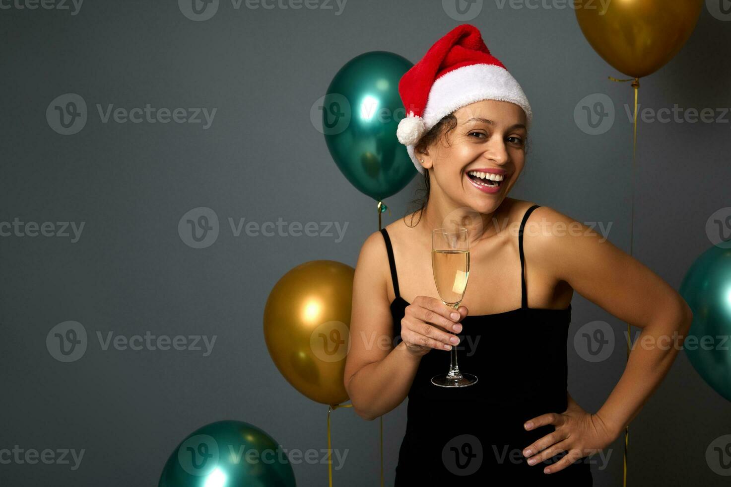 Beautiful young woman in Santa Claus hat and black evening dress rejoices and has fun at christmas party holding champagne flute isolated on gray background with shiny gold and green balloons photo