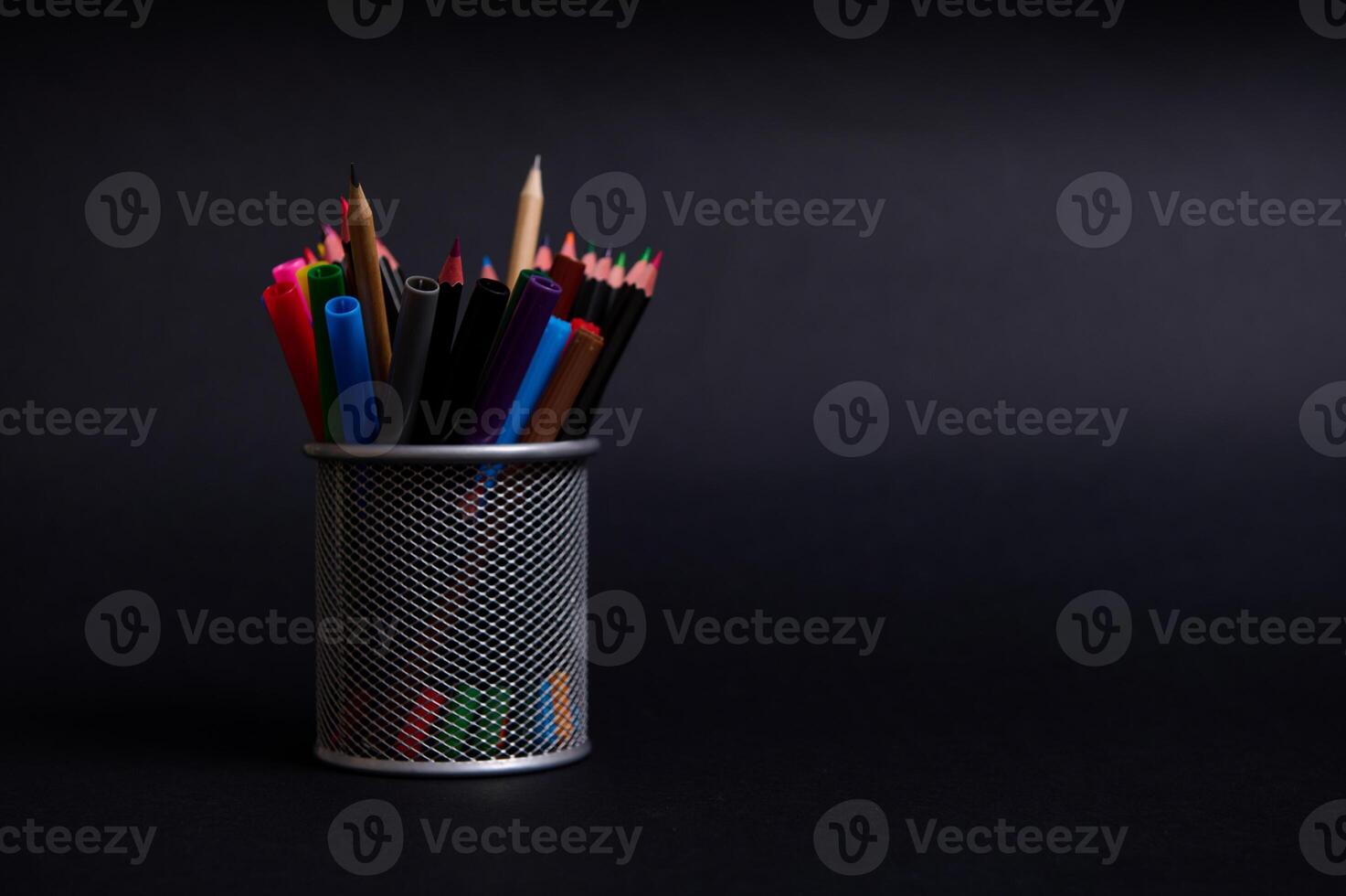 Stand for pens and pencils on the desktop on a black background with copy space. Close-up of a metal bucket with stationery. Holder with colored school supplies photo