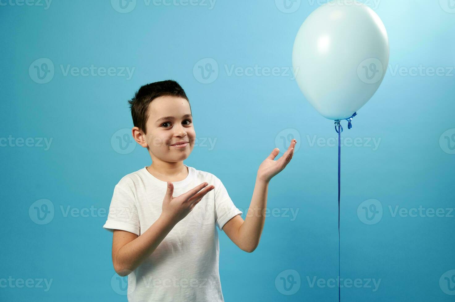 Handsome boy pointing on blue balloon and cute smiles looking at camera. Blue background with copy space photo