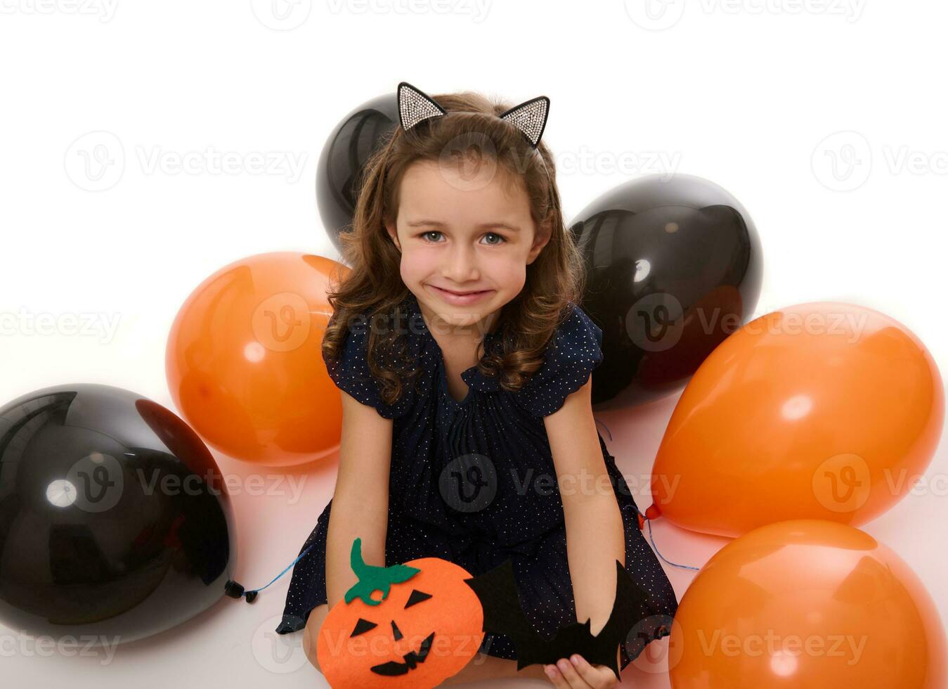 Beautiful girl child smiles with toothy smile , playing with homemade felt-cut pumpkin on the white background with lying down inflated black and orange colored balloons. Halloween concept, copy space photo