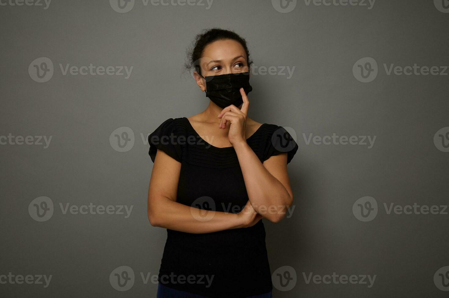 Isolated portrait on gray background of a woman in black medical mask with crossed arms looking thoughtfully to the side at a copy space for advertisement. Black Friday concept photo