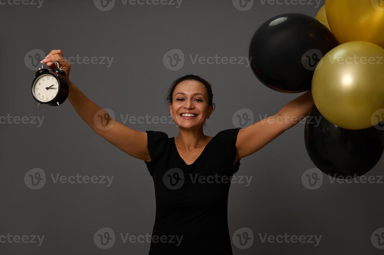 Cheerful woman in black attire raises her arms up holding gold and black inflated air balloons and a black alarm clock. Concept of Shopping at Black friday on gray wall background with copy ad space photo