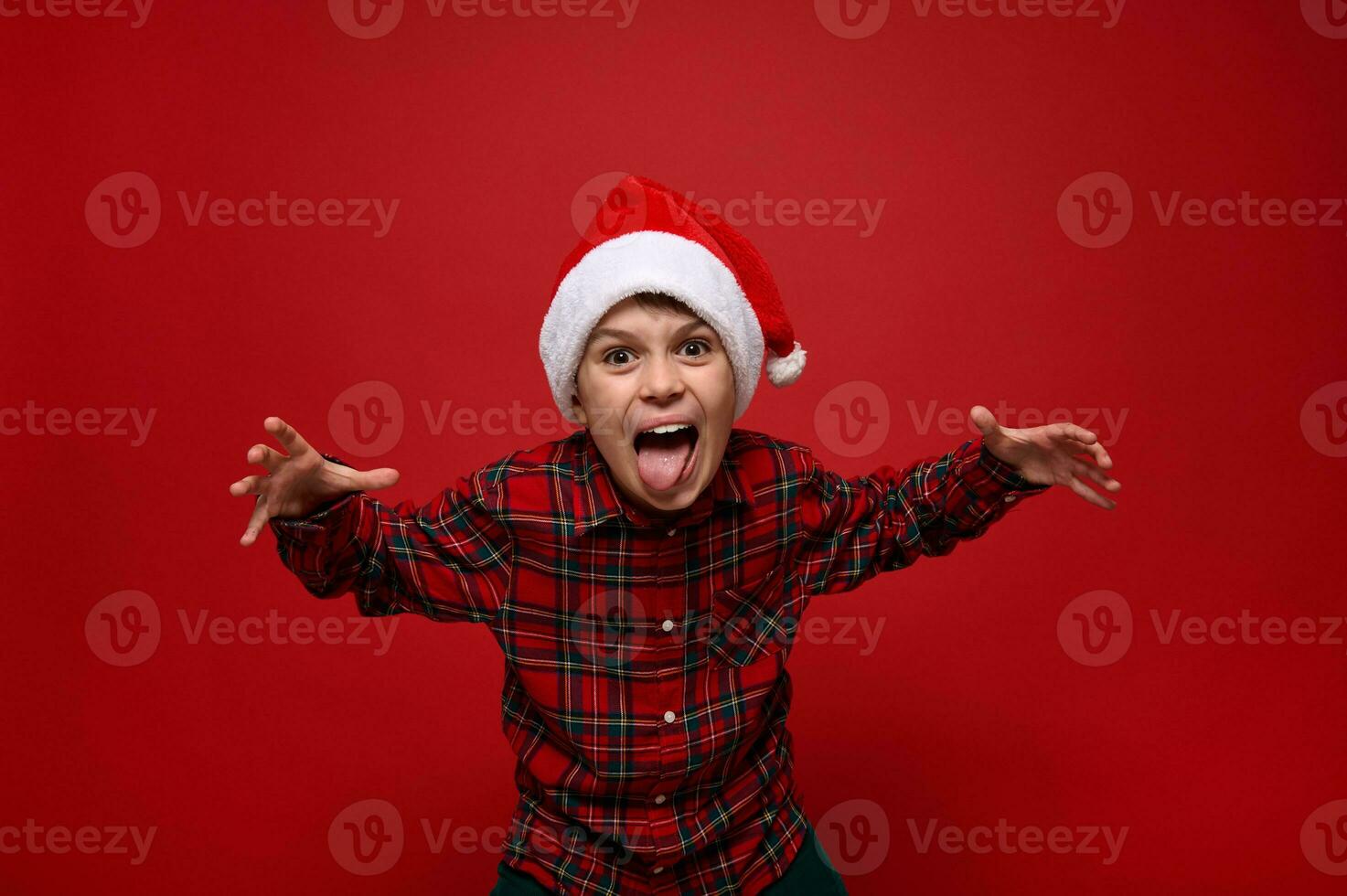 Funny cheerfule cute European preadolescent boy in plaid shirt and Santa Claus hat grimacing looking at camera posing against red colored background with copy space for New Year and Christmas ad photo