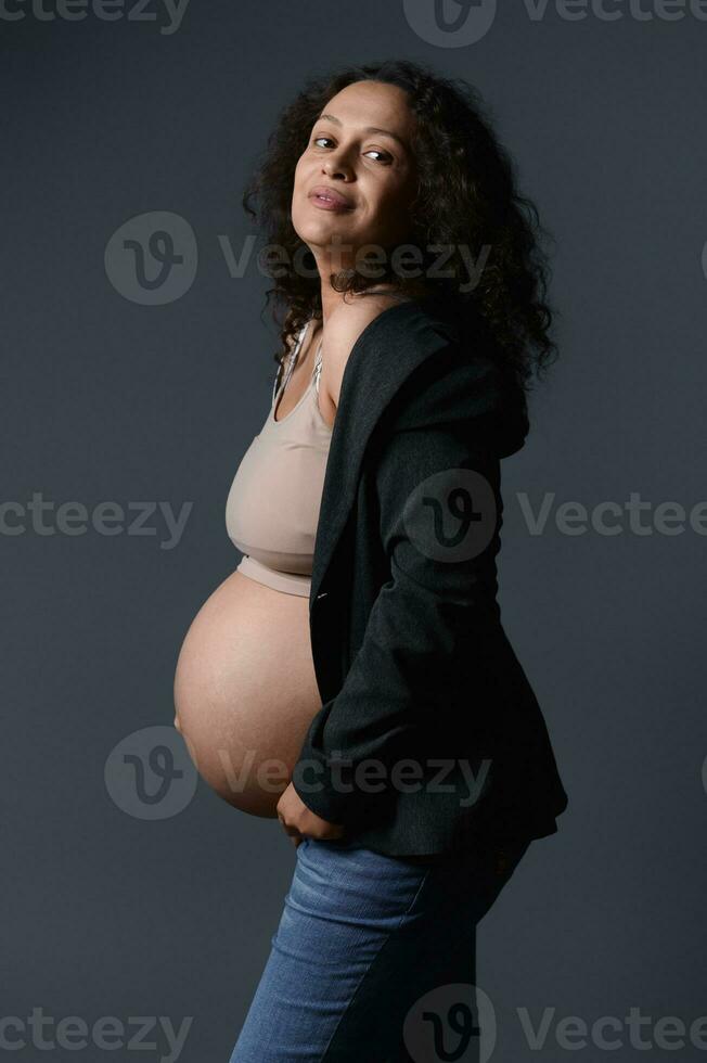 atractivo sonriente latín embarazada mujer en elegante chaqueta de sport, beige sostén y azul vaqueros, demostración su desnudo hermosa barriga foto