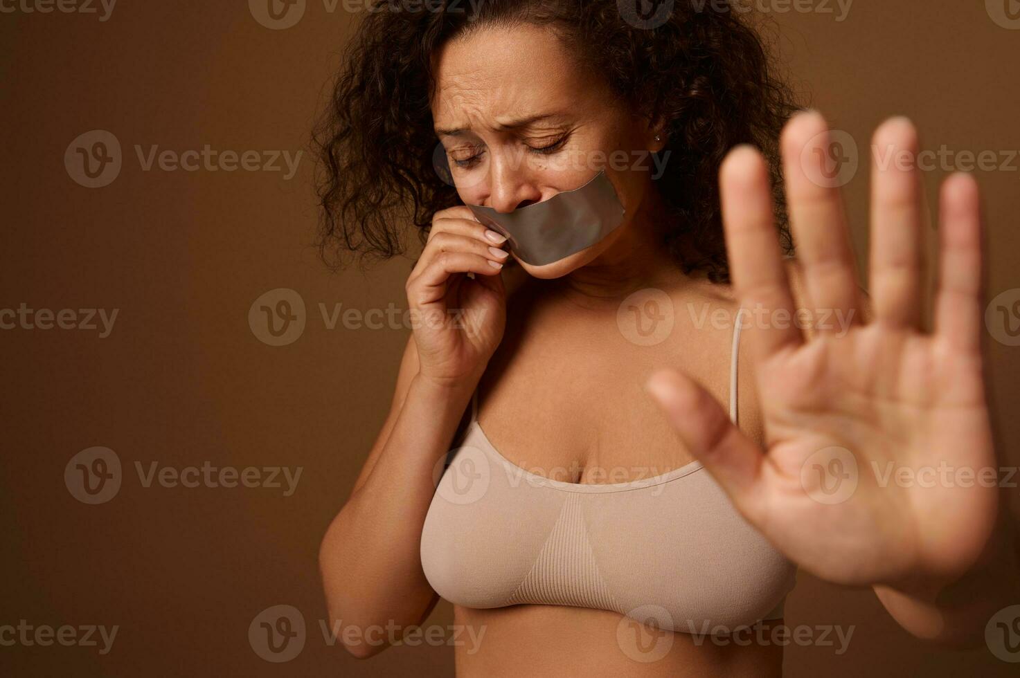 Desperately frightened crying woman with tears in her eyes, with sealed mouth shows STOP sign with her hand to camera, stands on dark beige background. Social concept of ending violence against women photo