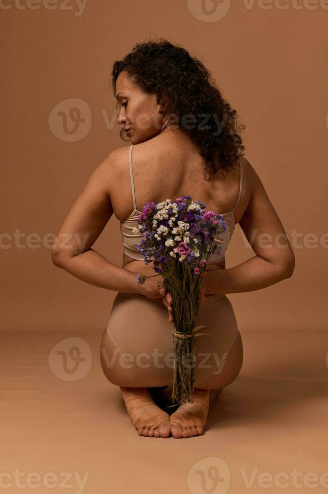 posterior ver de hermosa africano mujer en lencería, mirando a el lado, participación un ramo de flores de flores silvestres detrás su atrás, sentado en beige antecedentes. cuerpo positivo, auto-aceptación, feminidad concepto foto