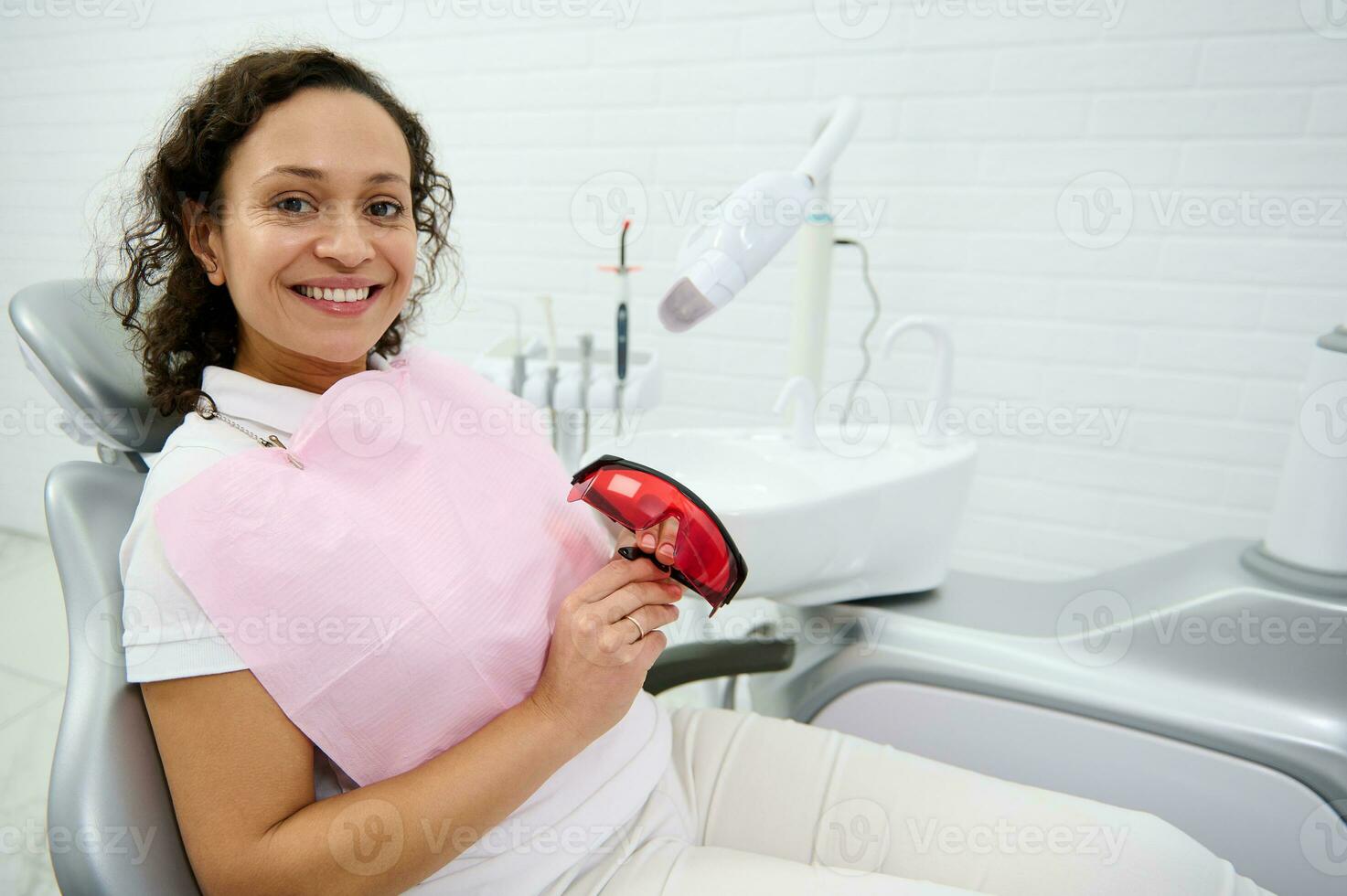 Cheerful African American woman sitting in dentists chair, holding UV protective goggles and smiles with beautiful toothy smile looking at camera after teeth whitening procedure in a dental clinic photo