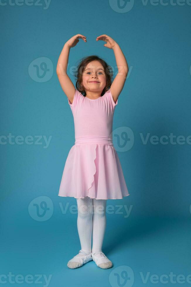linda sonriente bailarina en un rosado vestir realiza un actitud en un ballet danza mientras en pie con elevado manos en azul antecedentes foto