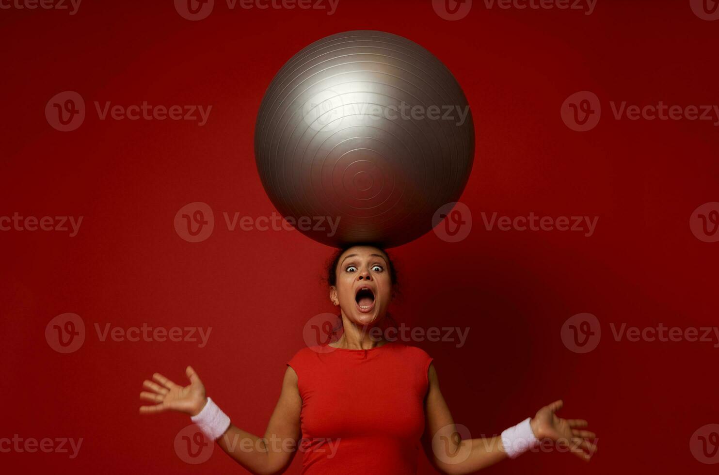 Astonished young sporty mixed race woman poses against red wall background with huge fitness ball on her head photo