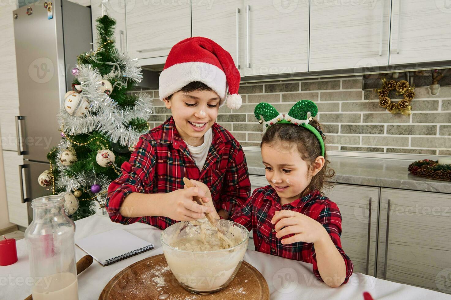 alegre Navidad y contento infancia concepto. linda pequeño niña con duende aro y su más viejo hermano- adorable caucásico preadolescente chico en Papa Noel sombrero, disfrutando Cocinando juntos a hogar cocina foto