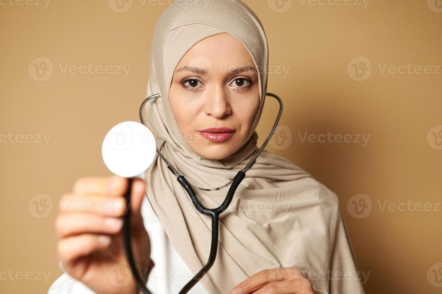 Confident portrait of a female muslim doctor wearing hijab and showing a stethoscope to the camera. Closeup on beige background with copy space. photo