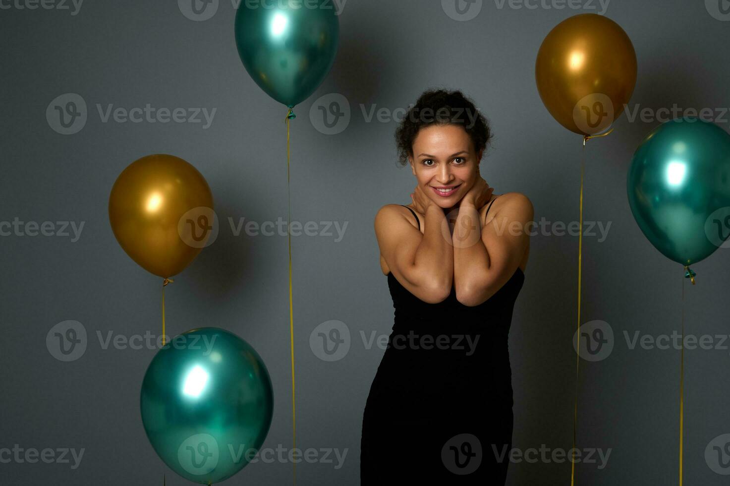 Portrait of happy pretty woman in evening black dress smiling with beautiful toothy smile posing against air balloons on gray wall background with copy space for Christmas, New Year, anniversary ad photo