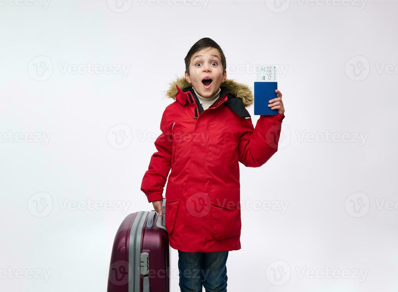 Happy school child, adorable handsome boy in bright red down jacket standing near a suitcase and holding his passport with ticket and boarding pass. Travel and winter holidays concept with copy space photo