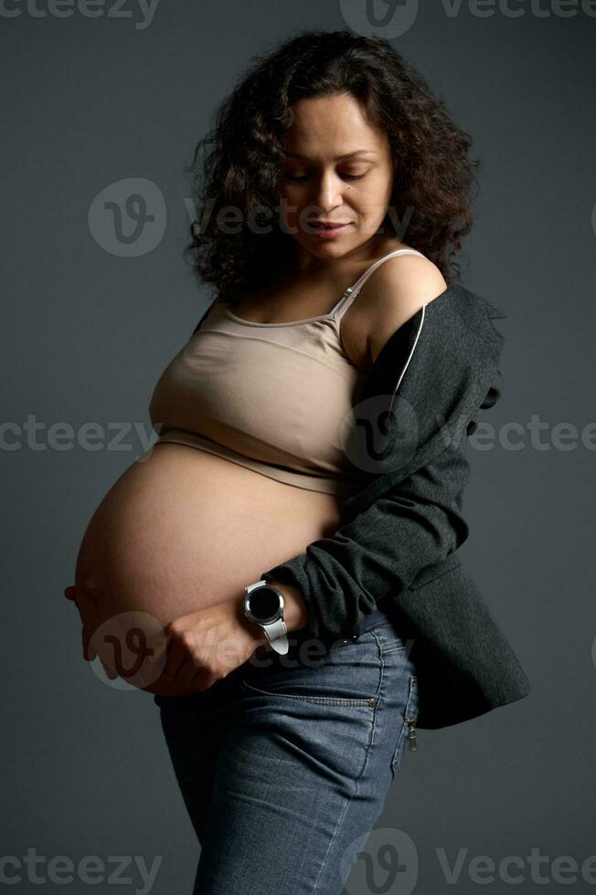 Curly haired Latin American pregnant woman, expectant mother touching her big belly in the ninth month of her pregnancy photo