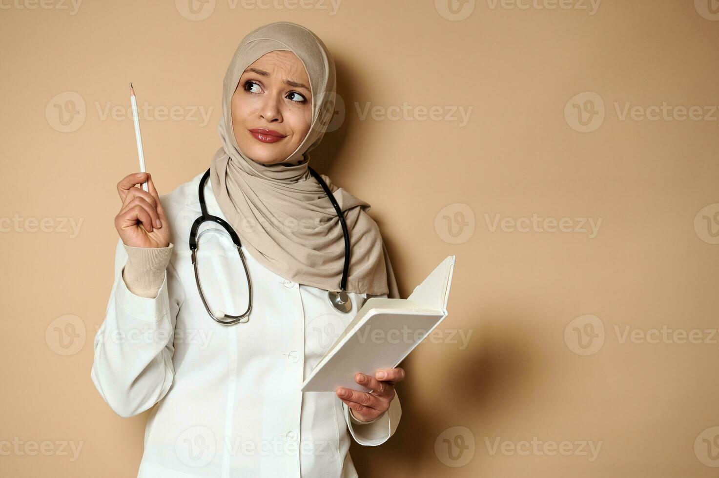 Attractive Arabic Muslim female doctor holds a diary and looks up thoughtfully, pointing with a pencil on a beige background with copy space. photo