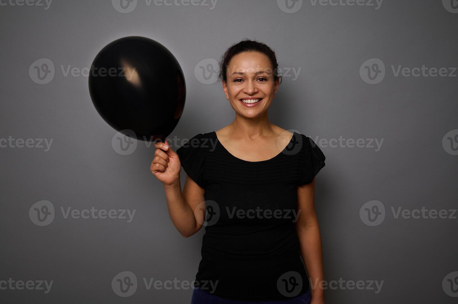 alegre africano americano mujer vestido en negro sonrisas mirando a cámara y poses en contra gris antecedentes con un negro de colores aire globo en mano. negro viernes concepto con Copiar anuncio espacio foto