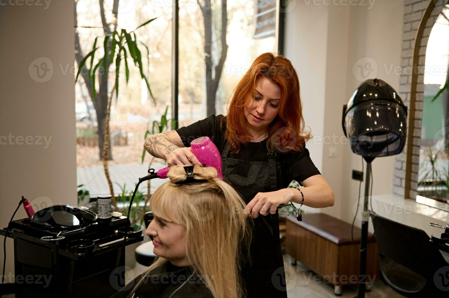 de moda hermosa Pelirrojo hembra peluquero utilizando un redondo peine y pelo secadora endereza rubia pelo a un cliente de un belleza salón, ejecutando su un elegante peinado. belleza industria concepto foto