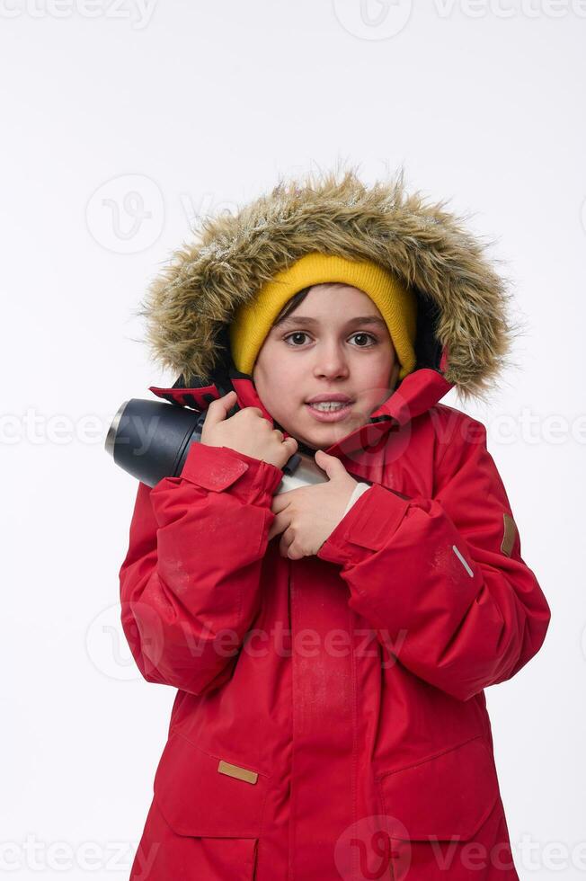 School-age traveler adventurer cute boy warming himself hugging thermos, shivering from cold looking at camera isolated on white background with copy space for ads. Winter leisure, holidays concept photo