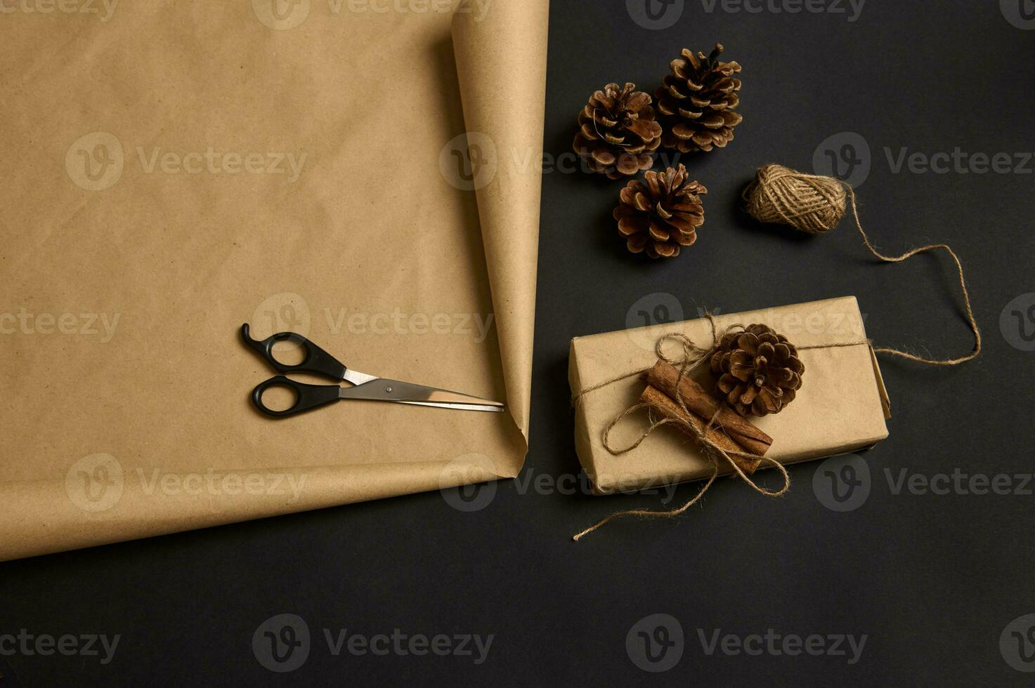 Top view of Christmas present decorated with pine cone and tied cinnamons, wooden numerals 2022, scissors and rope on brown craft wrapping gift paper on black surface with a copy space for ad photo