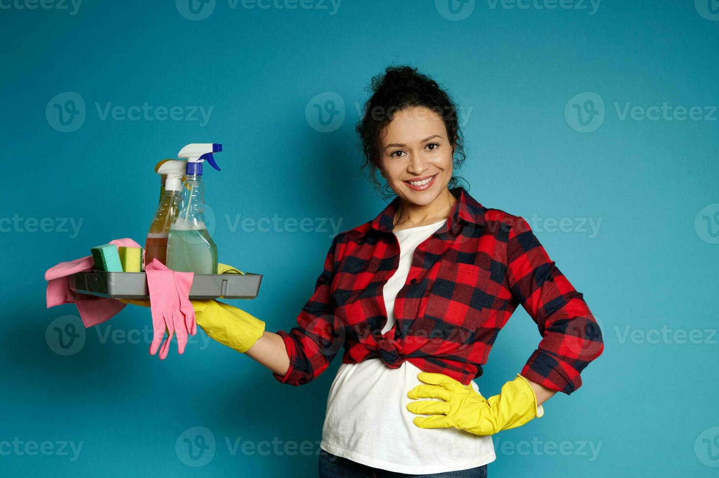 A charming brunette with curly hair, dressed in a white T-shirt, a red check shirt and yellow rubber gloves, holds a tray of cleaning supplies and cleaning tools. photo