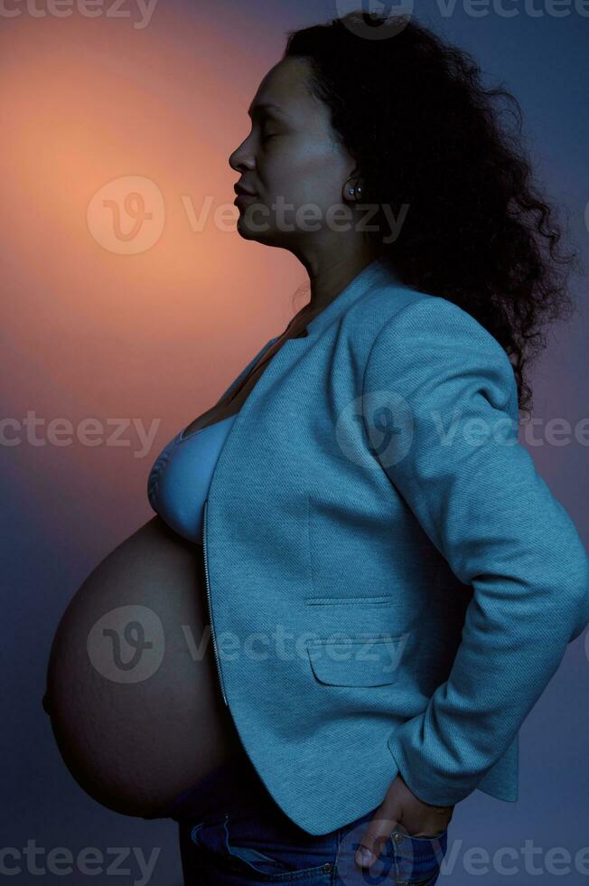 estudio retrato de un étnico embarazada mujer con grande barriga en el embarazo 36 semana, aislado en neón ligero color antecedentes foto